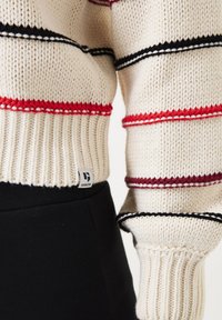 Suéter de punto grueso en crema con rayas horizontales rojas, negras y blancas. Presenta puños y cinturilla acanalados, con una etiqueta de marca visible.