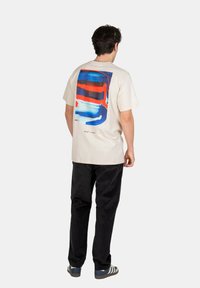 Mann in beigem T-Shirt mit abstraktem blauen und roten Grafik auf dem Rücken, schwarzen Hosen und blau gestreiften Sneakers, von hinten auf weißem Hintergrund.