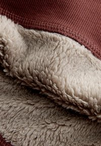 Brūnas ribotās auduma un maiga, bežīga mākslīgā kažokāda saskaras, izceļot gludu tekstūru un šuvju detaļas gar malu.