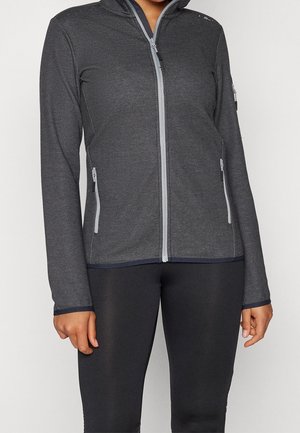 Femme portant une veste ajustée gris foncé zippée avec poches latérales zippées et des leggings noirs, debout devant un fond blanc.