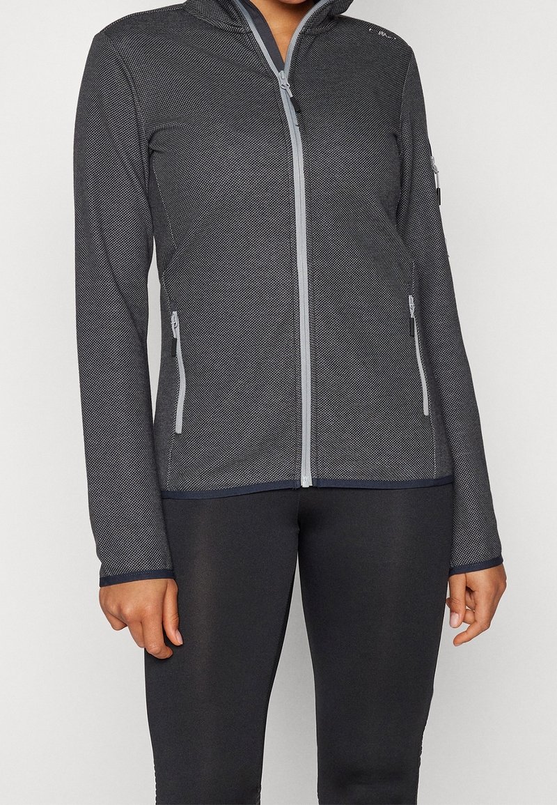 Femme portant une veste ajustée gris foncé zippée avec poches latérales zippées et des leggings noirs, debout devant un fond blanc.