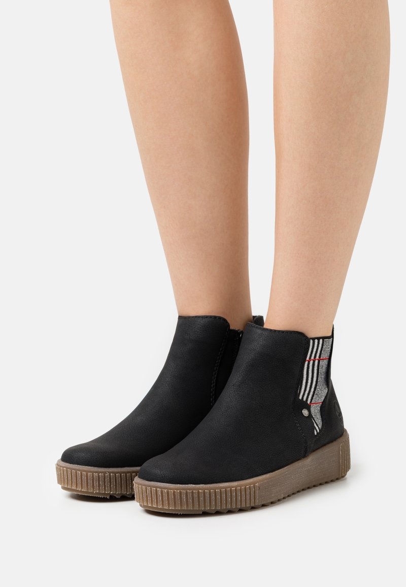 Bottines noires avec semelles épaisses marron et panneaux latéraux élastiques à carreaux, portées sur des jambes nues sur fond blanc.