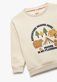 Beżowy sweter z okrągłą grafiką przedstawiającą drzewa, niedźwiedzia, ognisko oraz napisem "Przygoda zaczyna się tutaj. Natura to moje plac zabaw."