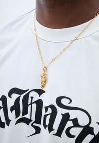 Colier din aur cu un pandantiv detaliat, purtat deasupra unei tricouri albe cu litere negre mari. Lanțul are zale ovale.