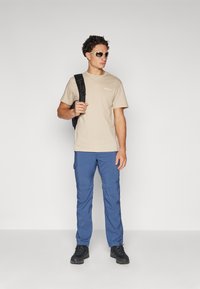 Tricou bej cu mânecă scurtă și logo, pantaloni cargo bleumarin cu buzunare, rucsac negru, ochelari de soare și pantofi de drumeție negri.