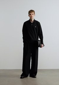 Μαύρο πόλο με μικρό λογότυπο, φαρδιές μαύρες παντελόνες και μαύρο τσαντάκι, φορεμένα με μαύρα παπούτσια. Απλός, μινιμαλιστικός στυλ μόδας.