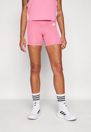 Person iført lyserøde sportsshorts og top, hvide sportssokker med sorte striber og hvide Adidas-sneakers, stående mod en ensfarvet baggrund.