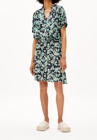 Robe fleurie bleu marine avec des motifs verts et roses, manches courtes, décolleté en V, taille élastique et jupe évasée. Associée à des chaussures slip-on beiges.