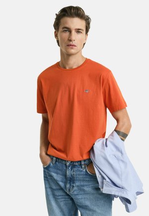 T-shirt di cotone arancione brillante con colletto rotondo e maniche corte, caratterizzato da un piccolo logo ricamato sul petto. Indossato con jeans azzurri.