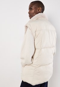 Puffer vest in lichtbeige met een pluizige kraag en gebreide mouwen. De achterkant heeft een iets langere zoom en een doorgestikt patroon.