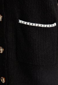 Czarny sweter dzianinowy z prążkowaną teksturą, z białym wykończeniem kieszeni i dużymi brązowymi guzikami z przodu.