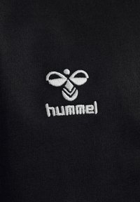 Schwarzer Stoff mit einer glatten Textur, der mit weißer Stickerei eines Logos verziert ist, das stilisierte Flügel und das Wort "hummel" darunter zeigt.