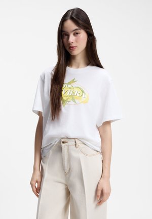 Ung kvinde med langt brunt hår iført en hvid T-shirt med grafik og beige højtaljede bukser, stående mod en ensfarvet baggrund.