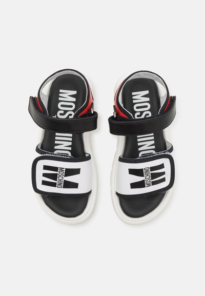 Las sandalias en blanco y negro cuentan con correas ajustables, una suela blanca gruesa y un logo negro en una correa blanca. Detalles rojos cerca del talón.
