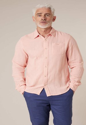 Homme aux cheveux blancs et à la barbe portant une chemise rose clair à boutons et un pantalon bleu foncé, debout les mains dans les poches devant un fond neutre.