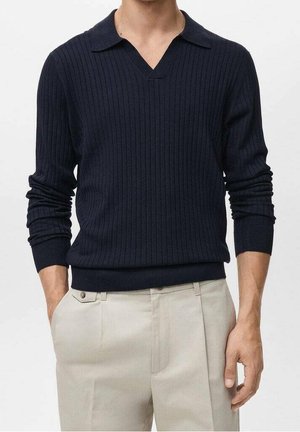 Mann trägt einen marineblauen gerippten Langarm-Strickpullover mit V-Ausschnitt und hellbeige Hose, die Hände locker seitlich.