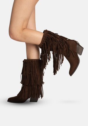 Jambes portant des bottes montantes en daim marron foncé avec détails à franges superposées et talons carrés empilés sur fond blanc.