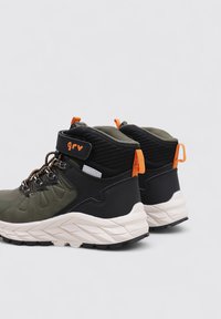 Chaussures de randonnée mi-hautes en vert olive et noir, en matériaux synthétiques, avec des lacets robustes, un logo orange et une semelle blanche texturée.