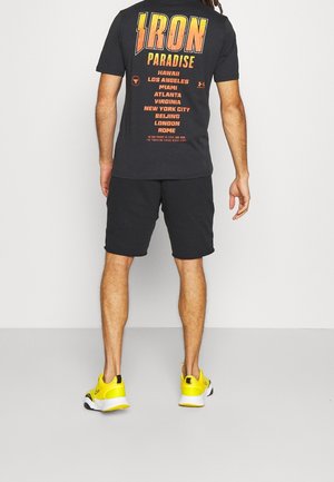 Mann trägt schwarze Shorts, ein schwarzes "IRON PARADISE" T-Shirt mit Stadtnamen und leuchtend gelbe Turnschuhe, steht mit dem Rücken zur Kamera vor hellem Hintergrund.