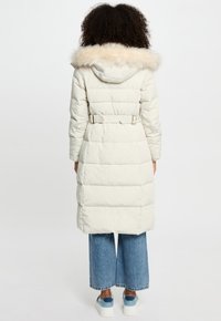 Giacca puffer beige chiaro con cappuccio in pelliccia sintetica, cintura in vita e motivo trapuntato verticale. Indossata sopra pantaloni in denim a gamba larga e sneakers.