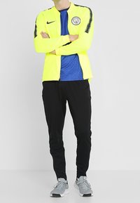 Bright yellow Nike jacket with black accents, a blue mesh shirt underneath, black pants, and gray sneakers. Design is athletic and fitted.