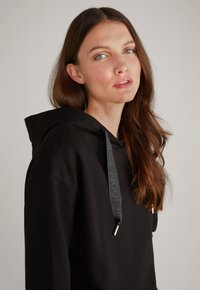Felpa nera con cappuccio e cordino che presenta il marchio "JOOP!", tessuto morbido, vestibilità rilassata e punte metalliche sul cordino.