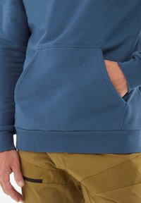 Niebieska bluza z kangurzą kieszenią, ściągaczem u dołu i przy mankietach; noszona z beżowymi spodniami z czarną kieszenią zapinaną na zamek po bokach.