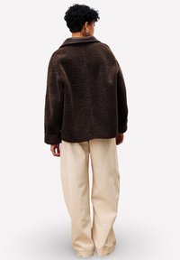 Personne avec des cheveux courts et bouclés portant une veste texturée marron foncé et un pantalon beige ample, se tenant de dos sur un fond blanc.