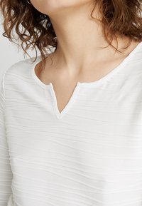 Top bianco con un motivo testurizzato a onde e un delicato scollo a V, realizzato in un tessuto morbido e leggero. Senza cuciture o parti metalliche visibili.