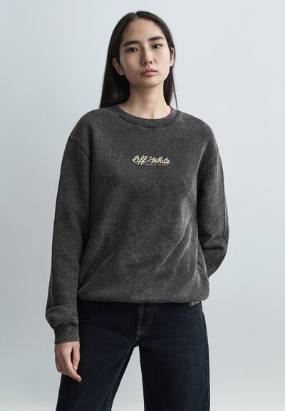 Sudadera de forro polar gris oscuro con cuello redondo, que presenta un logo dorado de "Off-White". Mangas largas con puños acanalados, combinada con jeans oscuros.