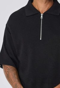 Polo noir à manches courtes avec une finition texturée et une fermeture à demi-glissière au niveau du cou. Présente un col plat et des poignets côtelés.