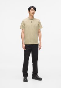 Béžová polo tričko s golierom a tromi gombíkmi, s malým logom na hrudi, spárované s čiernymi nohavicami a čiernymi topánkami.