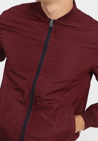 Chaqueta bomber burdeos de material ligero con cierre frontal, acentos en azul marino y dos bolsillos laterales. Cuello y puños de canalé.