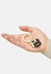 Gold metal keyring with a black and gold “B” logo charm, held in a hand. Smooth texture, rounded edges, lightweight design.