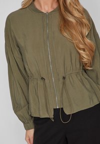 Jasnozielona, lekka kurtka z zamkiem z przodu, okrągłym kołnierzem i marszczoną talią. Posiada długie rękawy i regulowane sznurki u dołu.