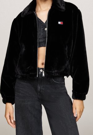 Person wearing a black fuzzy zip-up jacket with logo on chest, cropped black top, and high-waisted black jeans with subtle speckles.