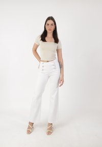 Ung kvinde iført en beige kortærmet top og højtaljede hvide bukser står med den ene hånd på hoften mod en ensfarvet hvid baggrund.