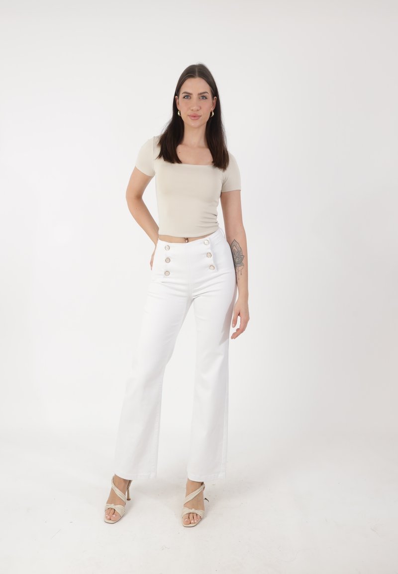 Mujer joven con una blusa de manga corta beige y pantalones blancos de cintura alta, de pie con una mano en la cadera frente a un fondo blanco liso.