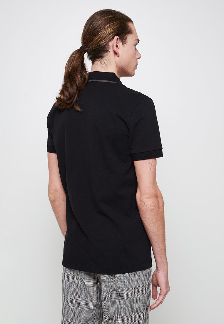 Homme avec de longs cheveux bruns en queue de cheval, portant une chemise noire à manches courtes et un pantalon à carreaux, se tenant de dos contre un fond blanc.