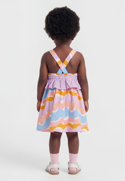 Mazais bērns ar cirtainiem matiem, tērpts pasteļtoņu kleitā ar krusteniski pārklātām lencītēm, rozā zeķēm un baltiem sandales, stāvot muguru pret skatītāju.