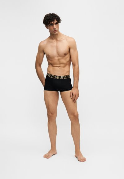 Homme torse nu portant un boxer noir avec une ceinture à motif clé grecque dorée, debout pieds nus sur un fond clair uni.