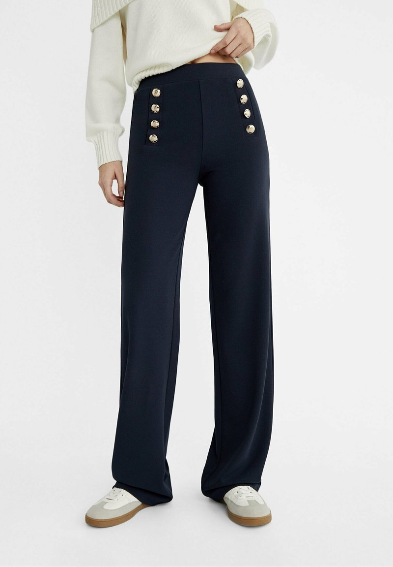 Pantalons larges bleu marine à taille haute, ornés de six boutons dorés à l'avant. Tissu doux et design lisse et structuré. Porté avec un pull blanc.