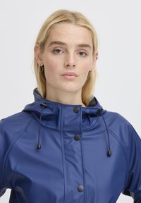 Giacca impermeabile blu con colletto alto, cappuccio regolabile con cordino, bottoni neri e una texture liscia e lucida.