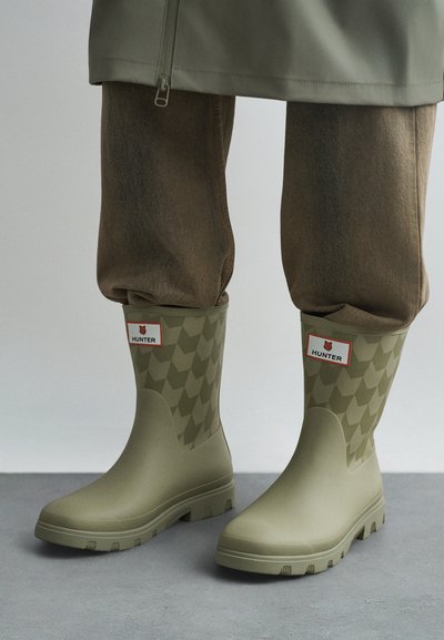 Zelena kaki Hunter škornja za dež in geometrijski vzorec, oblečena z rjavimi hlačami, ki so zataknjene notri, stoji na sivi tleh.