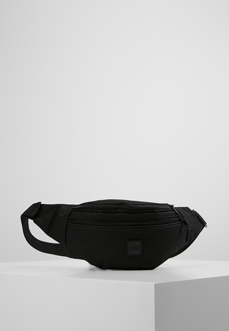Sac banane en tissu noir avec deux compartiments zippés et une sangle réglable, posé sur une plateforme blanche contre un fond gris uni.