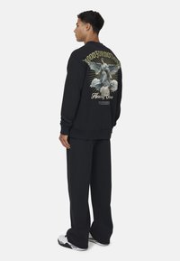 Felpa nera con una stampa grafica sulla schiena che raffigura una figura alata e testo. Abbinata a pantaloni scuri e sneakers bianche.