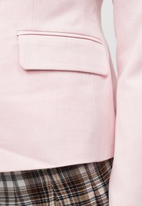 Κοντινό πλάνο στον φάκελο της τσέπης ενός ανοιχτόροζ σακακιού πάνω σε ύφασμα καρό φούστας σε καφέ, μαύρο και λευκό χρώμα, αναδεικνύοντας την υφή του υφάσματος και τις λεπτομέρειες του ενδύματος.