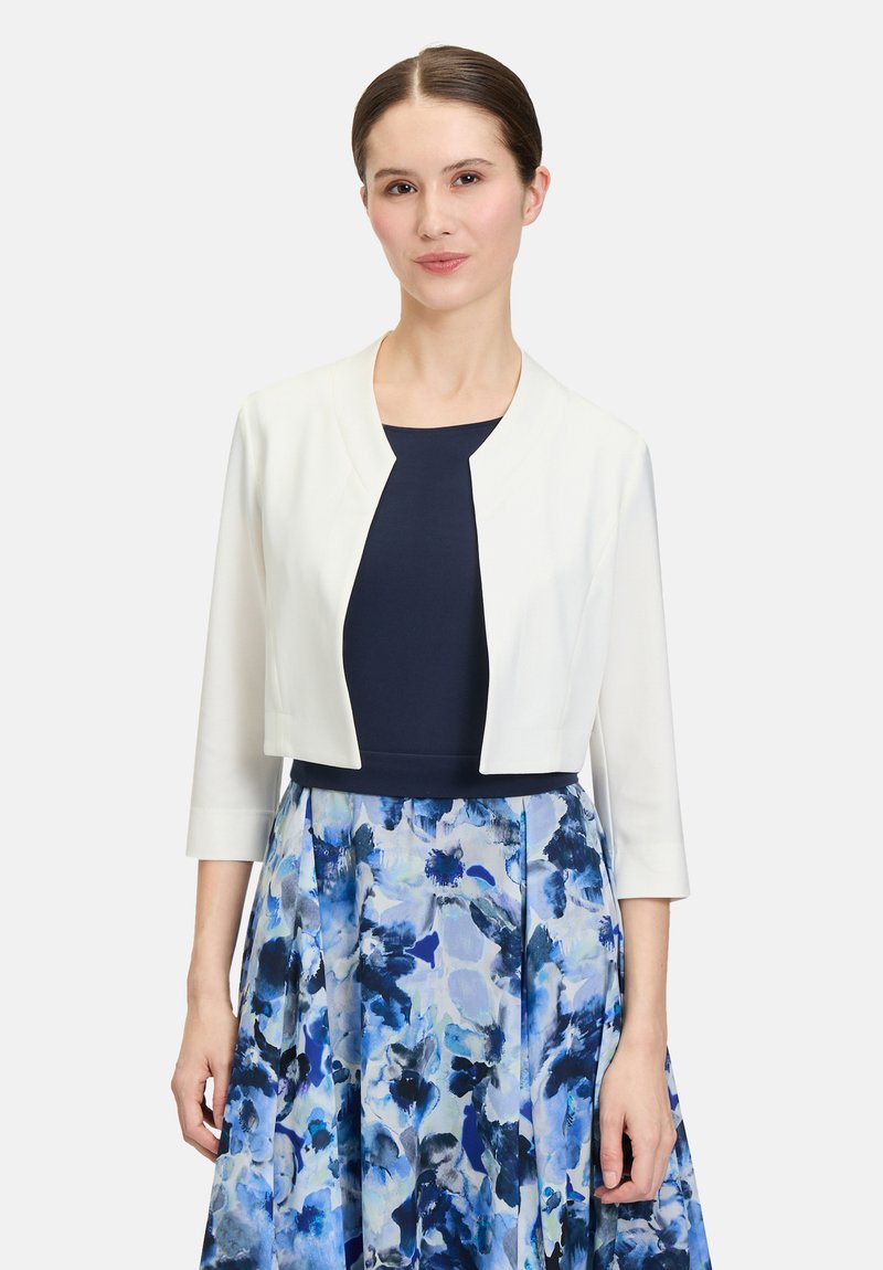 Witte cropped jas met een ronde halslijn, gecombineerd met een marineblauw top en een meerkleurige blauwe bloemenrok. Soepele textuur, 3/4 mouwen.