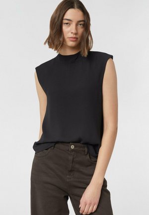 Frau mit schulterlangem braunem Haar, die ein ärmelloses schwarzes Oberteil und dunkelbraune Hosen trägt, vor einem einfarbigen weißen Hintergrund.