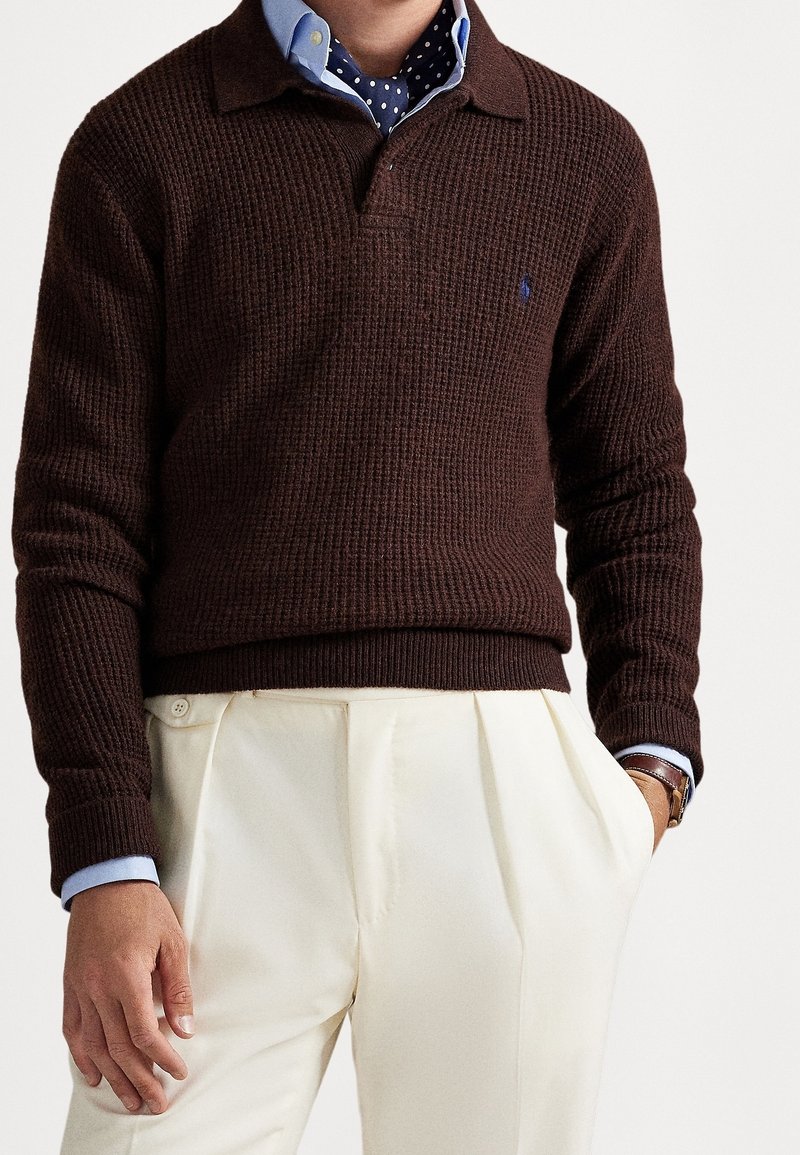 Personne portant un pull en maille marron foncé sur une chemise bleue à pois, avec un pantalon plissé beige clair, main dans la poche et montre-bracelet visible.